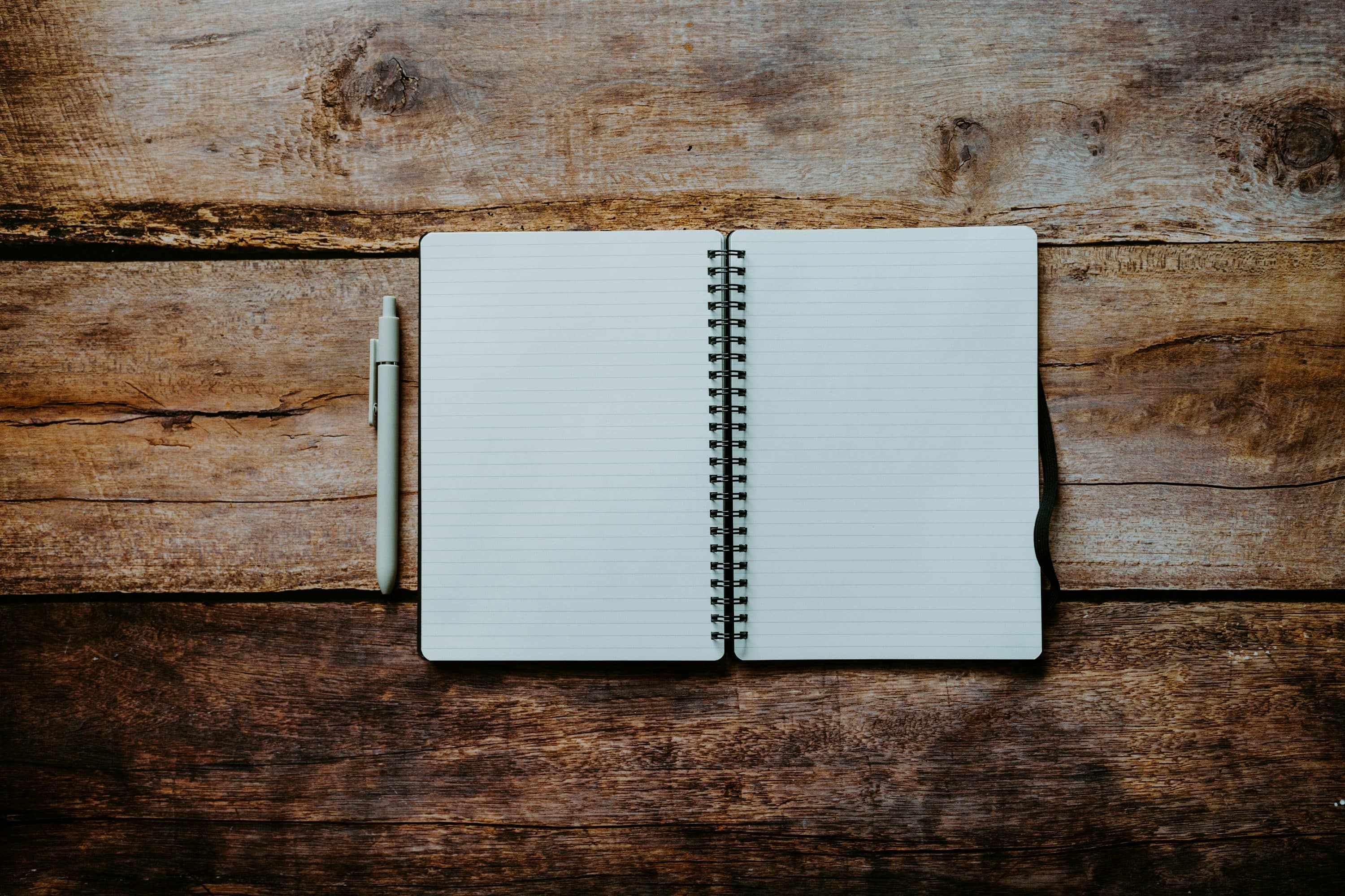 An open blank lined spiral notebook with a pale pen beside it on a rough wooden table.
